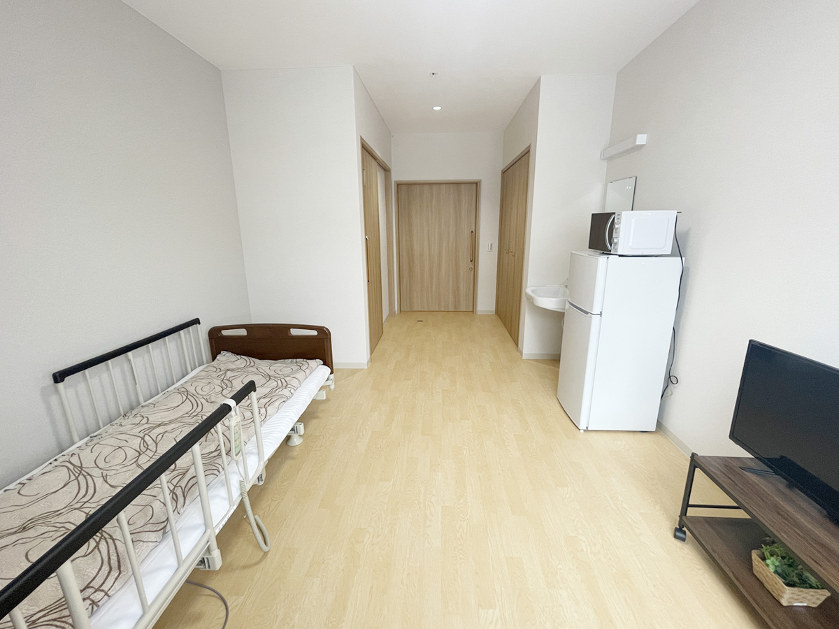 介護施設の個室。ベッド、冷蔵庫、テレビ、トイレを備えた居住スペース。